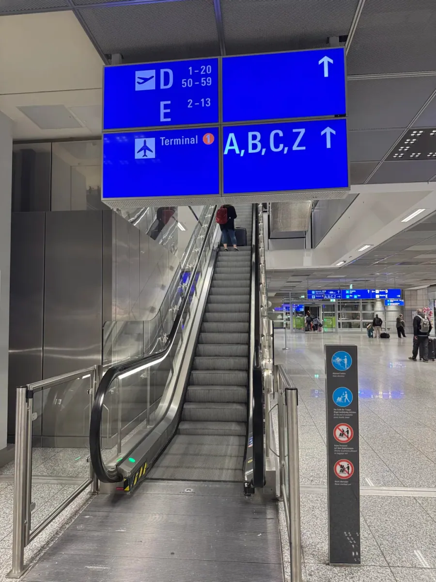 Escalator inside Terminal 2 up to the SkyLine level, with blue signs pointing to "D, E" and "A, B, C, Z" overhead