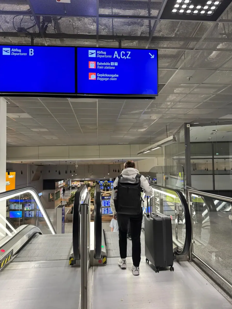 An escalator down inside Terminal 1, with signs overhead reading Bahnhöfe/Train stations and Gepäckausgabe/Baggage claim, and a traveller with a case descending