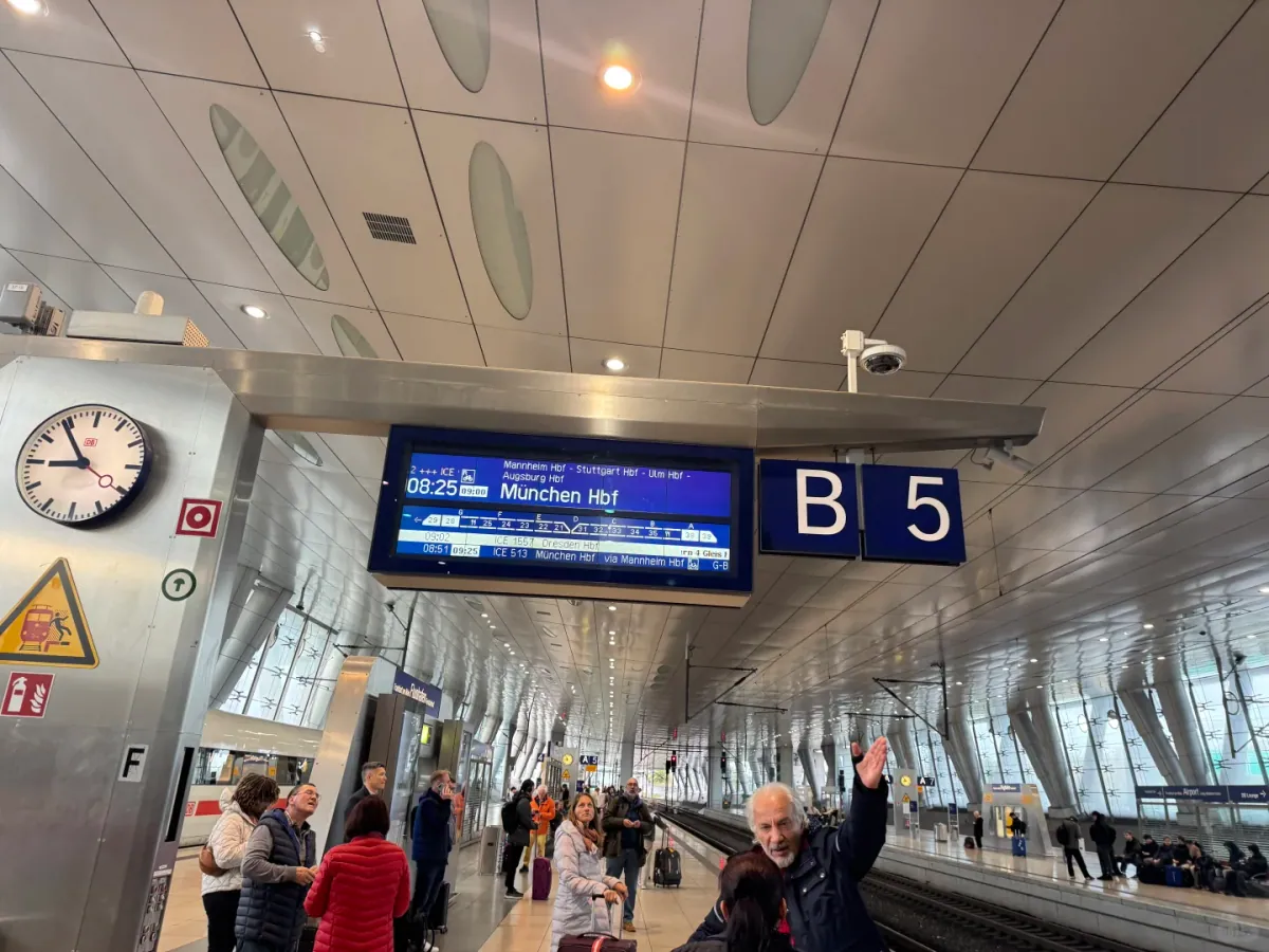 A blue platform sign reading B 5 at the Fernbahnhof, beside a display for the 08:25 ICE to München Hbf and a station clock, with passengers waiting