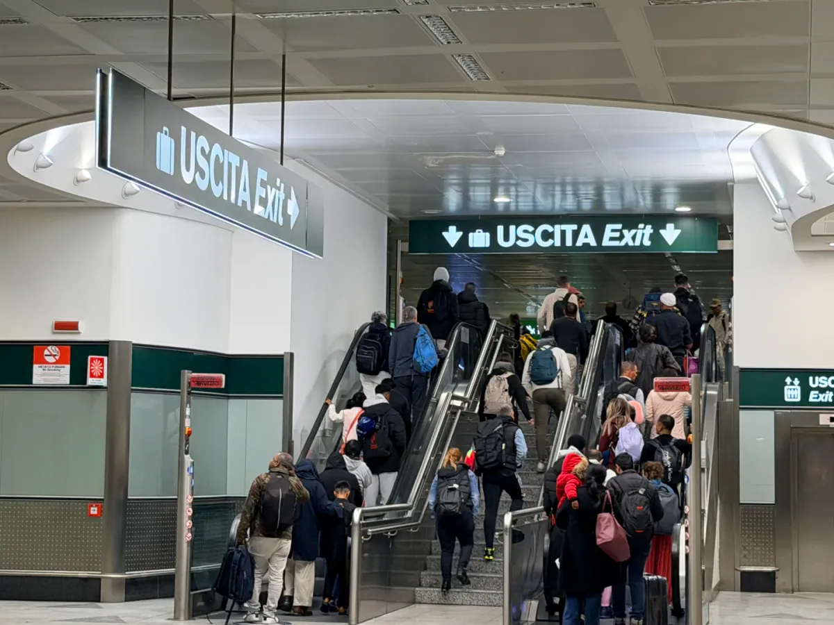 Passengers with luggage riding the escalator up towards the immigration hall at Milan Malpensa, under a black sign reading USCITA Exit