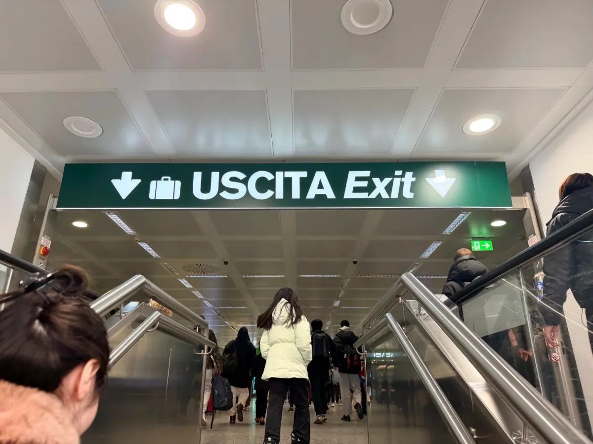Looking up from the escalator: a green sign reading USCITA Exit with a suitcase icon, with passengers walking towards the end of the corridor