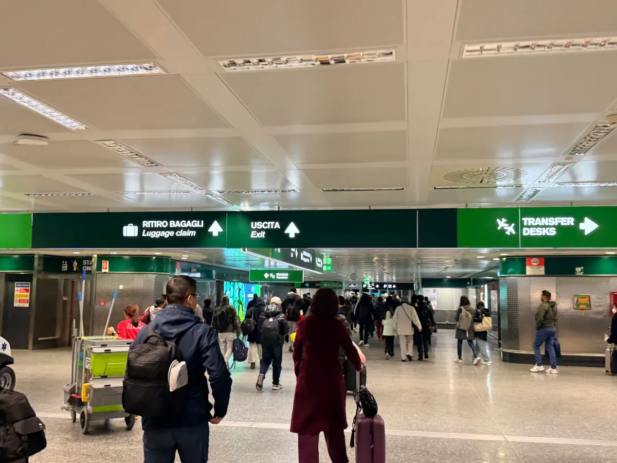 Green overhead signs in the Malpensa arrivals hall: RITIRO BAGAGLI / Luggage claim on the left, USCITA / Exit in the middle and TRANSFER DESKS on the right, with passengers walking through