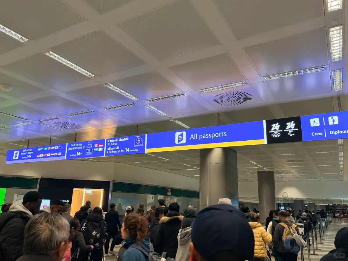 A long blue overhead sign across the corridor showing Self service passport control, All passports and Crew / Diplomatic, with the Milano Cortina 2026 Winter Olympics logo at the right-hand end and passengers queueing below