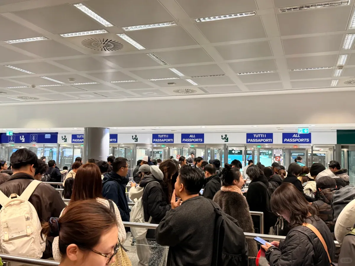 Passengers queueing at a row of TUTTI I PASSAPORTI / ALL PASSPORTS glass booths in the border hall, the snake barrier filled end to end