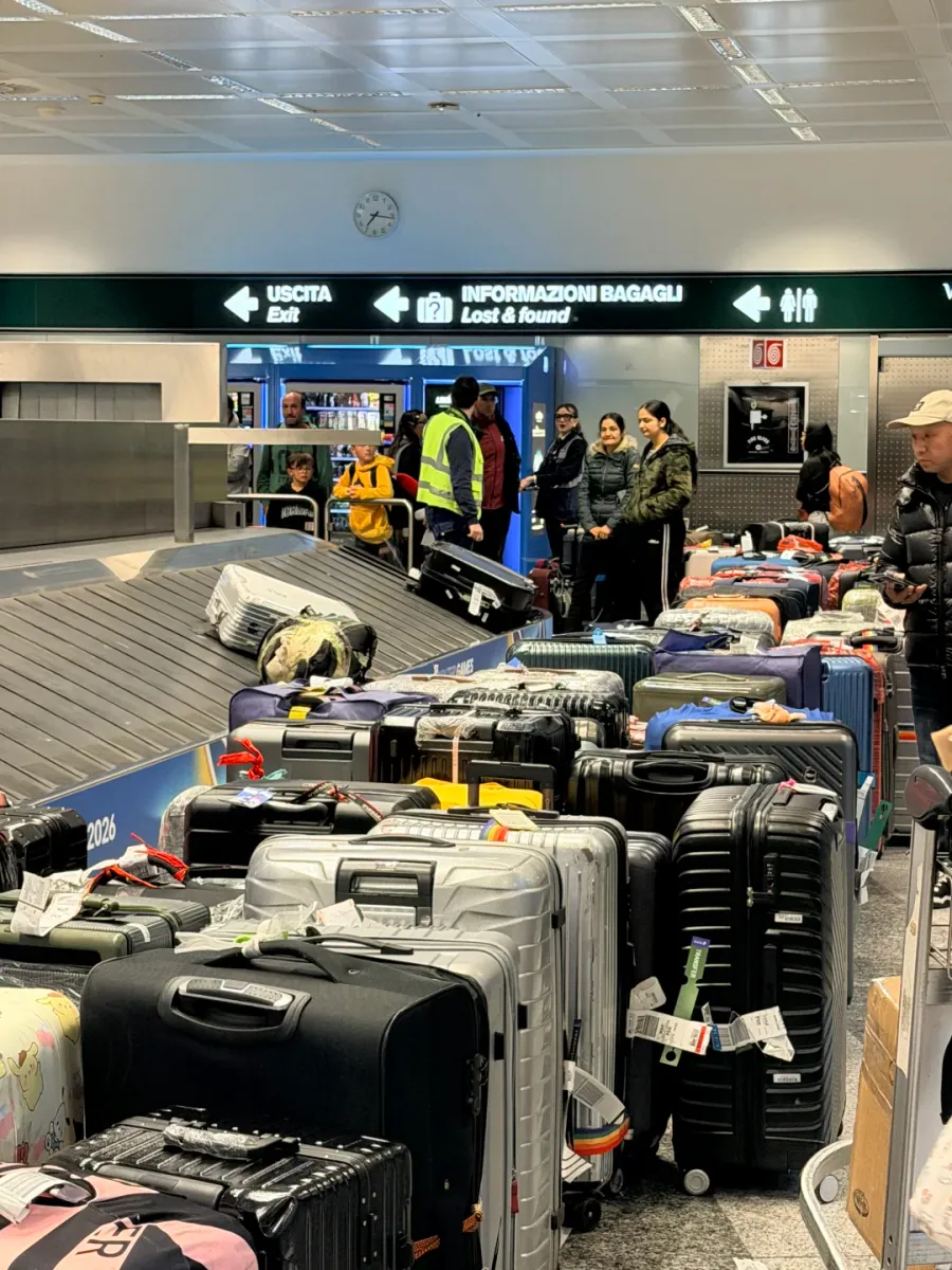 The baggage hall: a green sign reading USCITA Exit and INFORMAZIONI BAGAGLI / Lost & found, suitcases piled on the belt and on the floor beside it, with a worker in a fluorescent yellow vest standing by