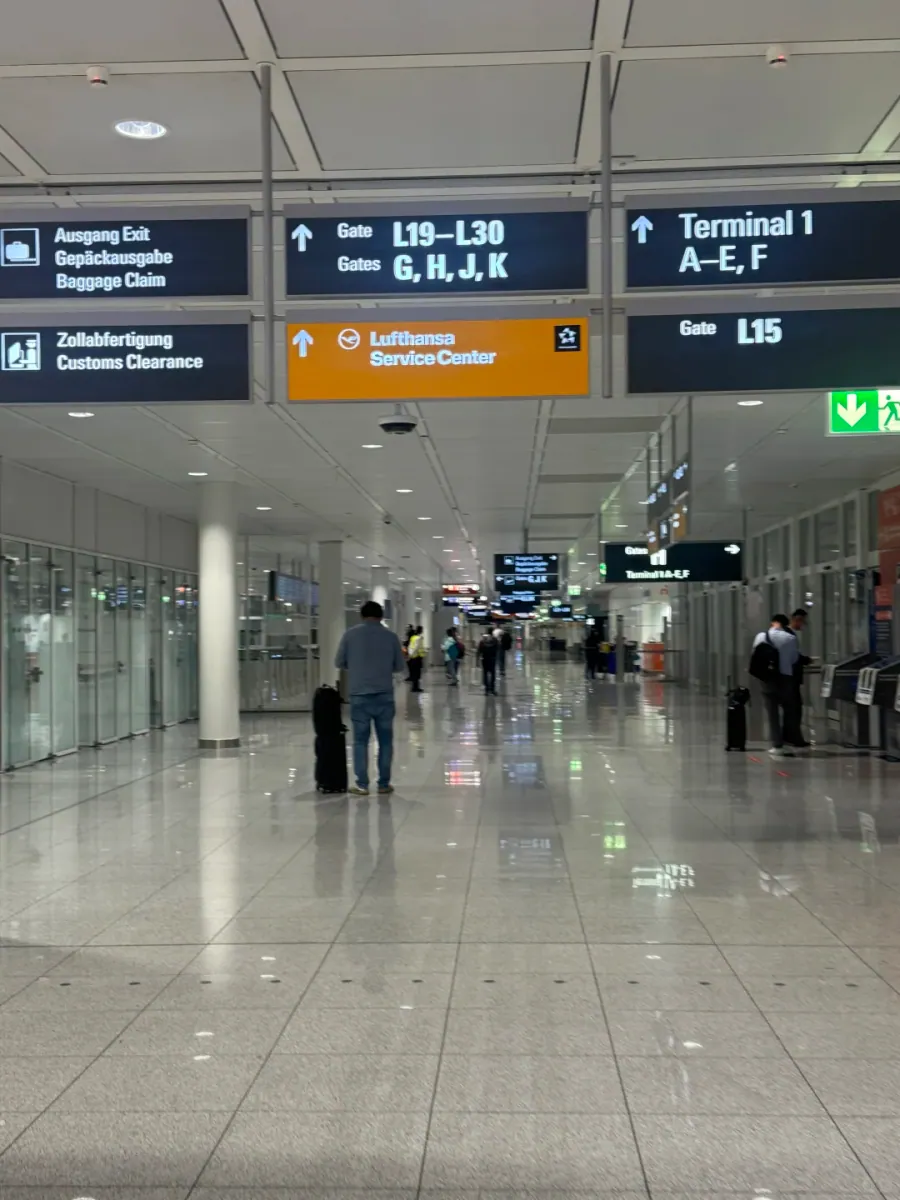 Dark blue overhead signs in a Munich Airport transfer corridor: Exit and baggage claim to the left, Gates G, H, J, K and L19–L30 straight ahead, Terminal 1 A–E, F to the right