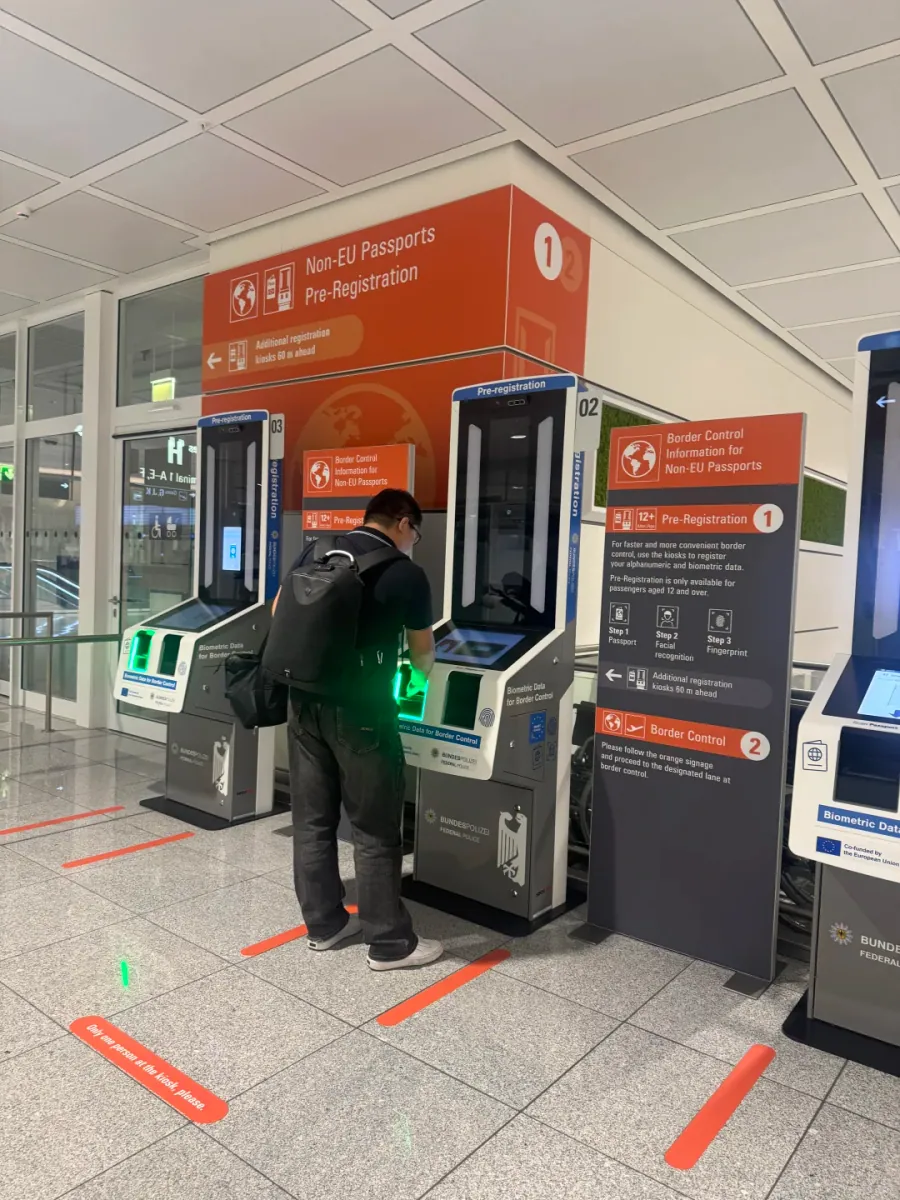 Two orange EES pre-registration kiosks under an orange canopy, a traveller with a backpack operating one, and a sign listing the three steps: passport, facial recognition, fingerprints