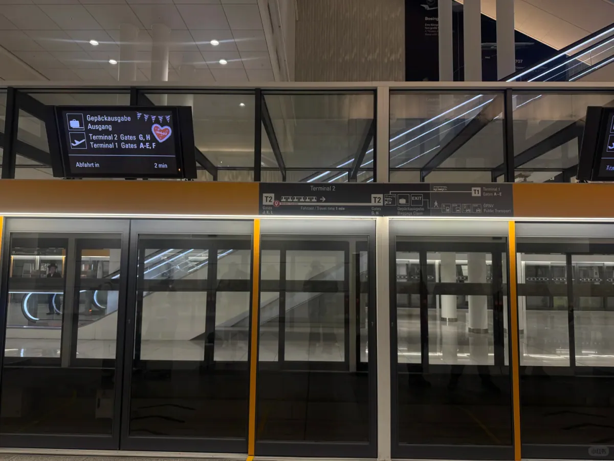 Glass platform screen doors with yellow frames at the people-mover station, the display above showing Terminal 2 gates G and H, Terminal 1 gates A–E and F, and departure in 2 minutes