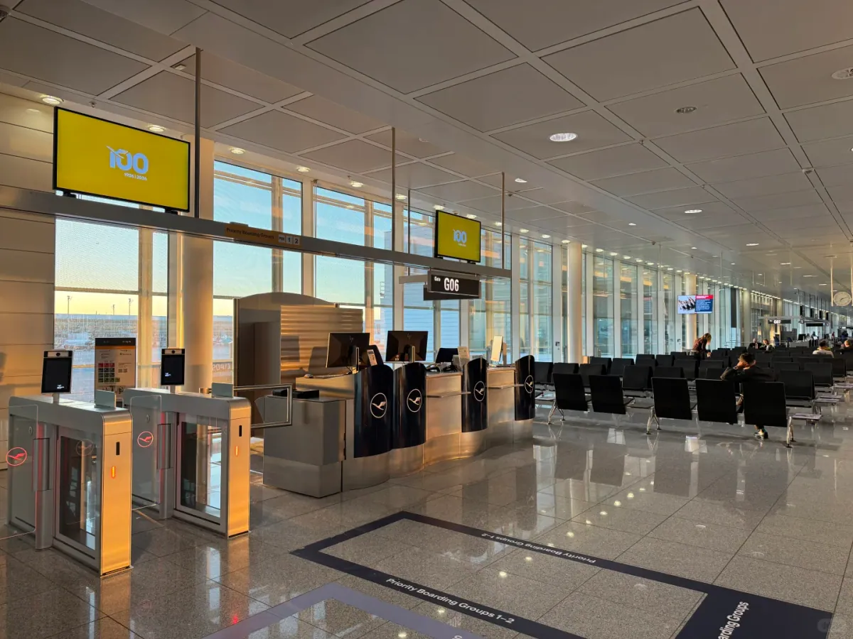 The gate concourse in the G area: self-service boarding gates under yellow screens, black leather seating, a black sign reading G06, and the apron in the early morning light