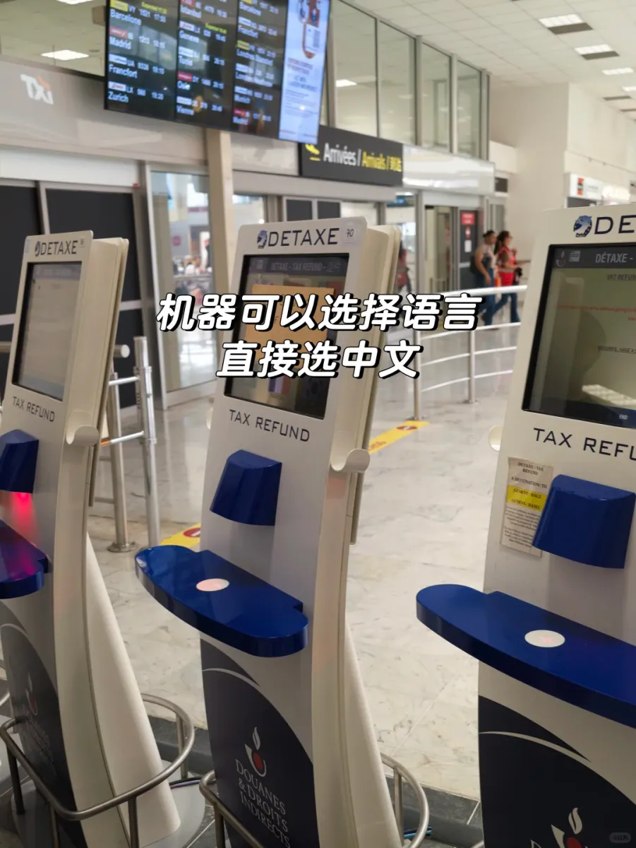 Several DETAXE self-service kiosks side by side in the Terminal 1 hall, white bodies with blue scanner hoods, in front of a flight information screen and an Arrivées / Arrivals sign