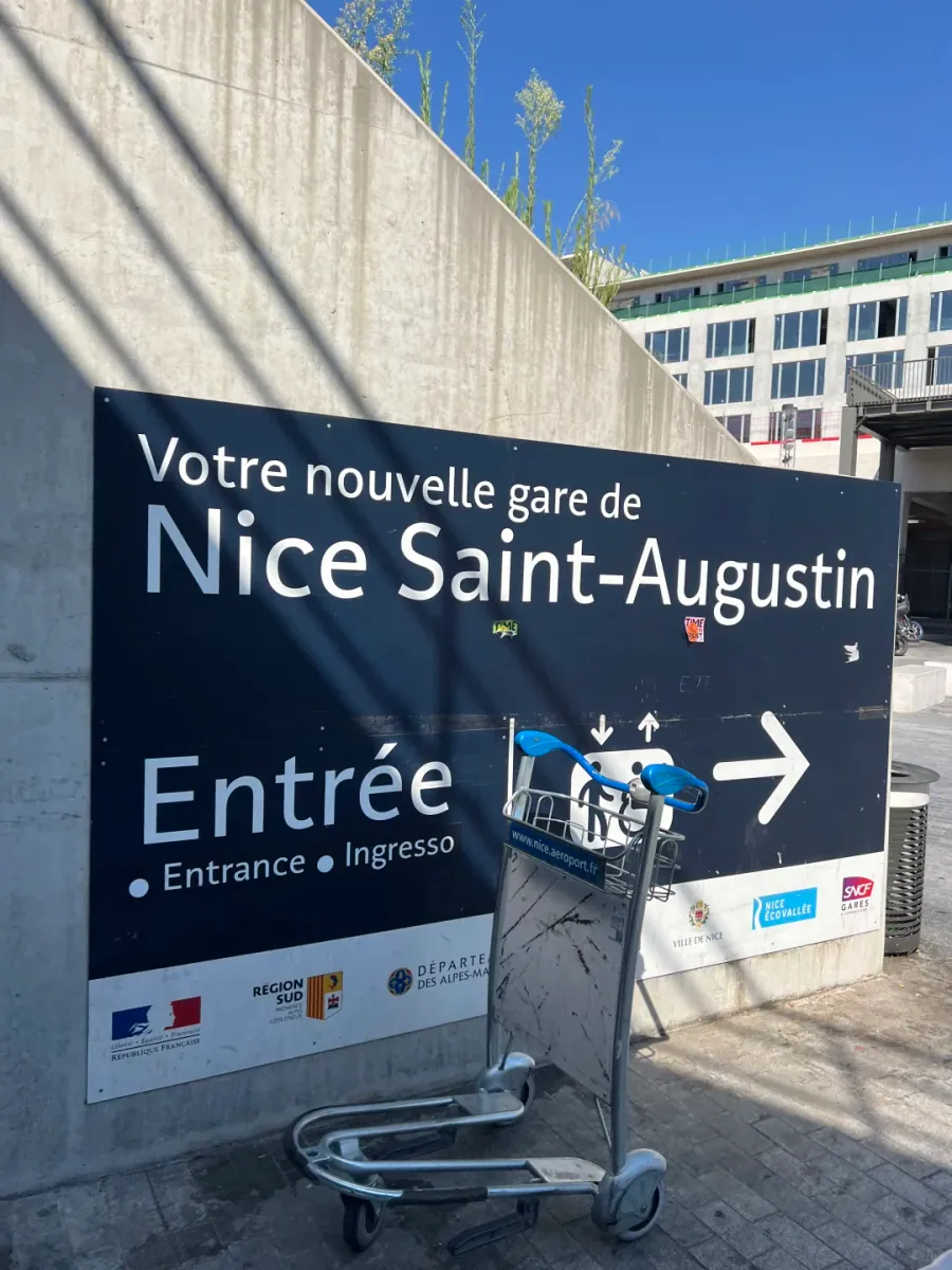 A dark blue board outside Saint-Augustin station reading "Votre nouvelle gare de Nice Saint-Augustin" and "Entrée", with an airport luggage trolley in front