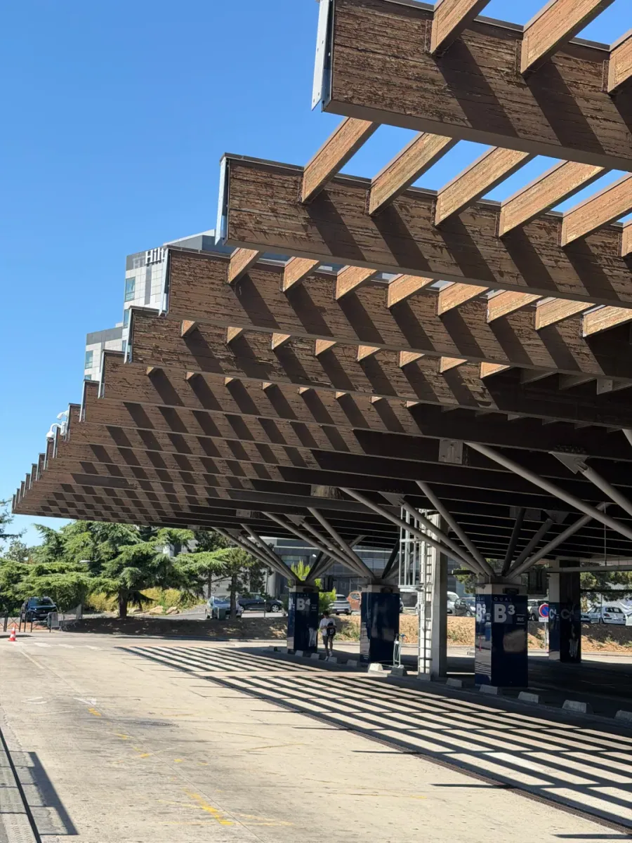 Coach stand B3 under a slatted timber canopy on steel columns, with a hotel tower and parked cars in the distance