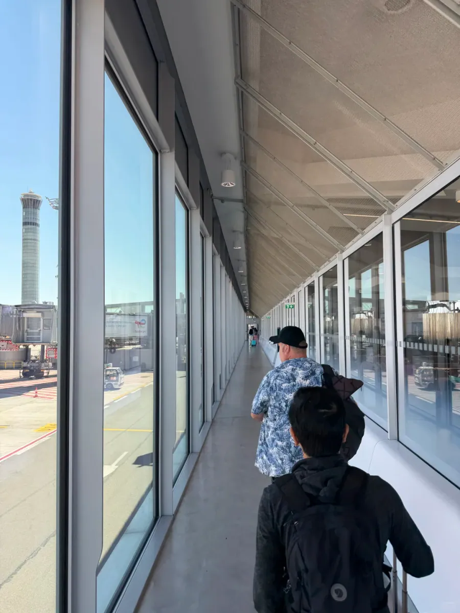 Travellers walking down a glass corridor, with the apron, airbridges and the airport control tower visible through the windows on the left