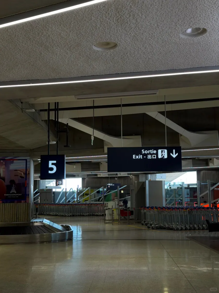 The baggage hall: a carousel beside belt 5, rows of luggage trolleys, and a hanging "Sortie / Exit / 出口" sign with a downward arrow