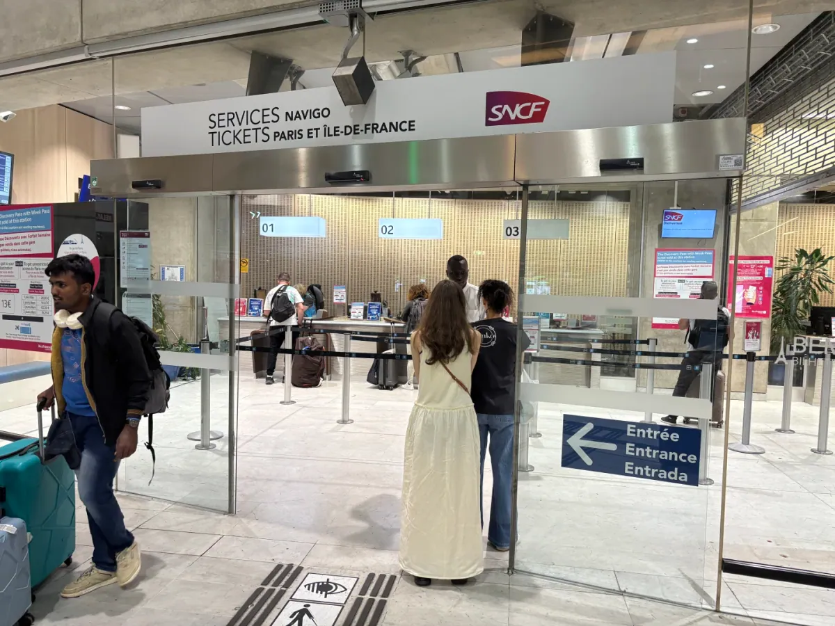 The SNCF ticket office inside the CDG station: "Services Navigo / Tickets Paris et Île-de-France" above the glass doors, people queueing at counters 01 to 03, and an "Entrée / Entrance" sign at lower right
