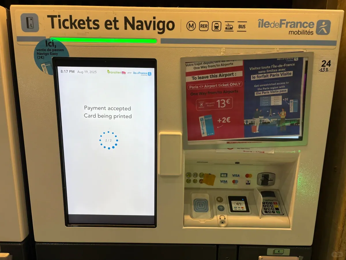 Close-up of a Tickets et Navigo machine: the screen shows "Payment accepted / Card being printed", with the "Paris ↔ Airport ticket 13€ +2€" poster beside it