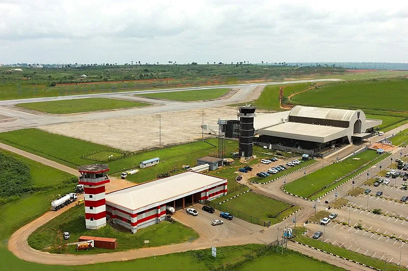 Luftaufnahme des Asaba International Airport: die bogenförmigen Terminaldächer und der dunkle Kontrollturm neben der Startbahn, im Vordergrund die rot-weiße Feuerwache und ein Tankwagen.