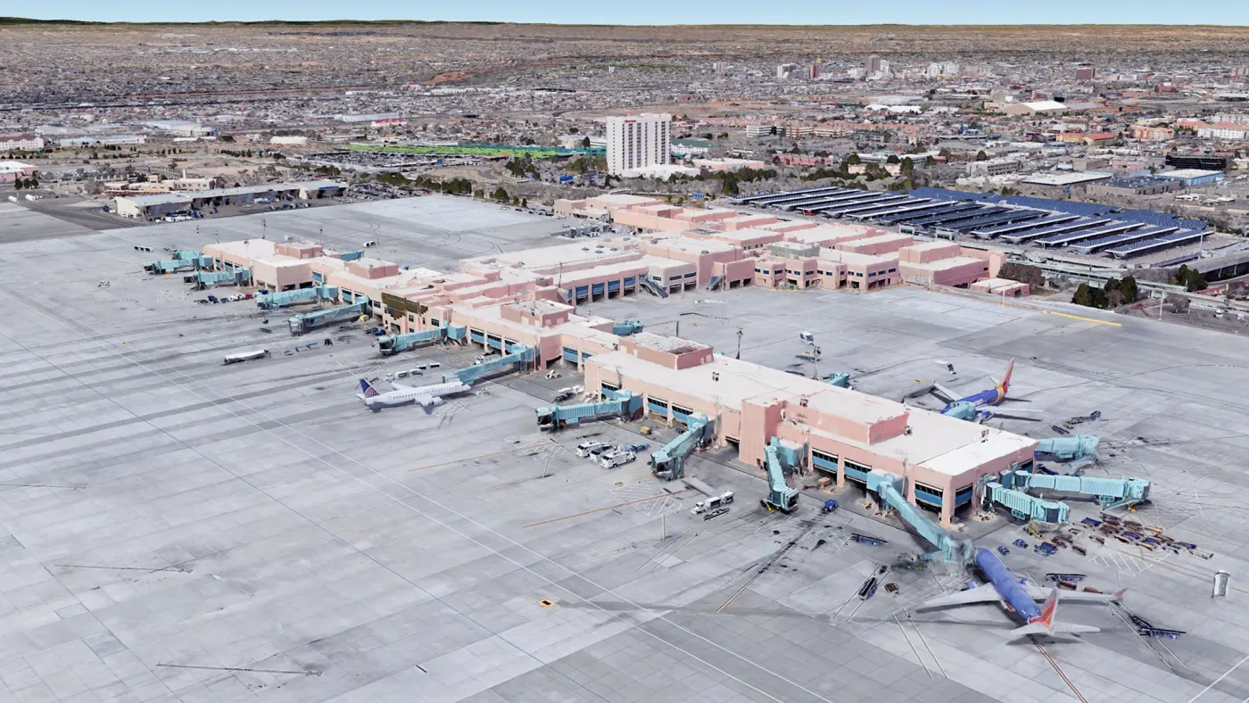 Luftaufnahme von Terminal und Vorfeld: rosa Concourses im Pueblo-Stil, über Fluggastbrücken mit dem Vorfeld verbunden, Flugzeuge an den Gates, dahinter Parkdecks und die Stadt.