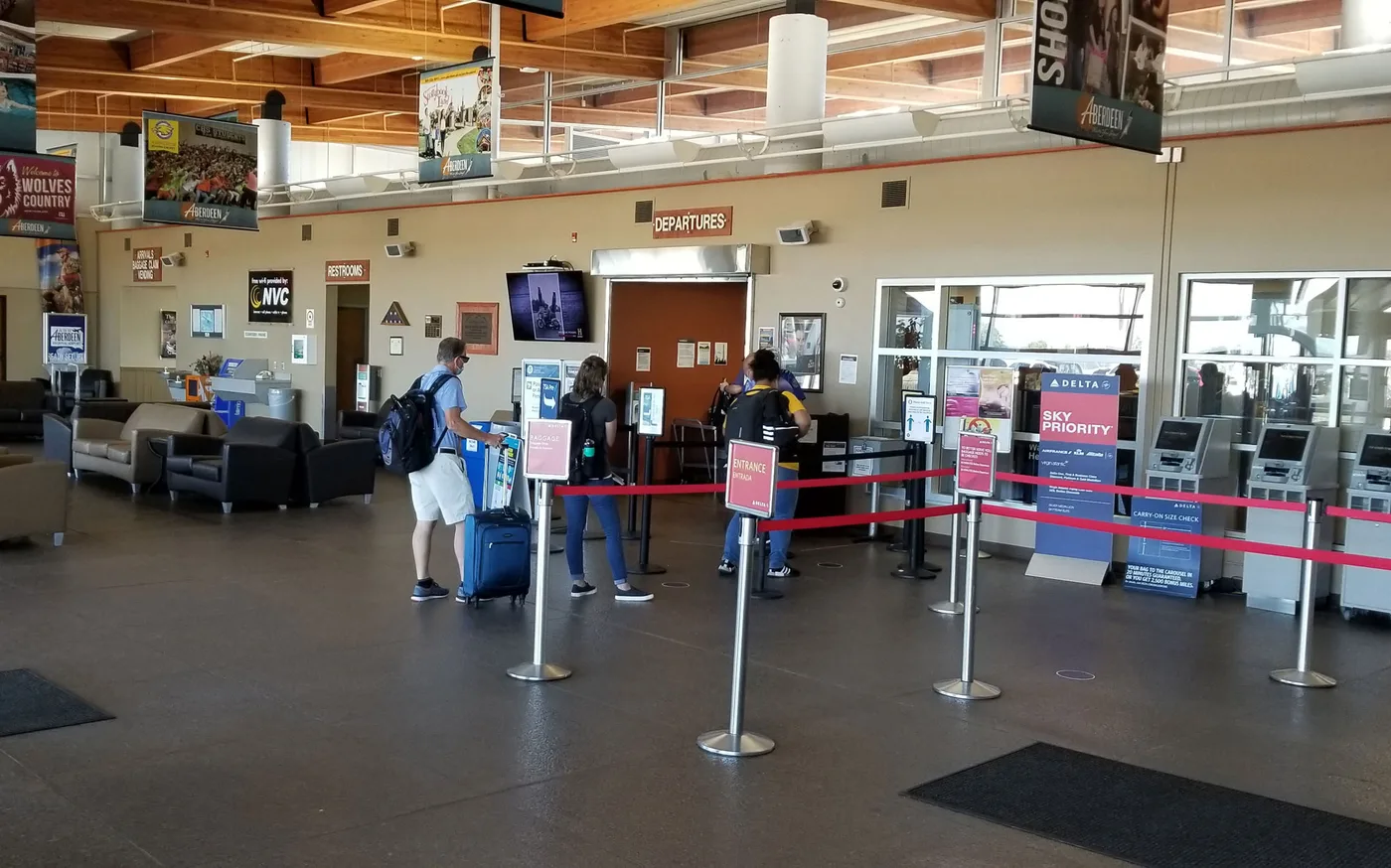Die Warteschlange in Schlangenlinien vor der Tür „DEPARTURES“, mit Delta-Selbstbedienungsterminals und Check-in-Schaltern rechts.