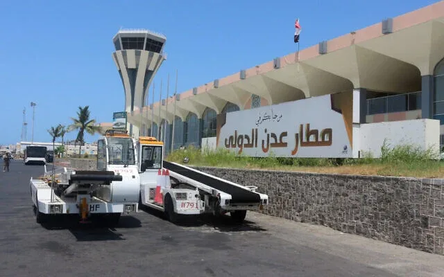 Terminal und Kontrollturm des Aden International Airport – der Name des Flughafens auf Arabisch an der Wand, mit zwei Pushback-Schleppern davor.
