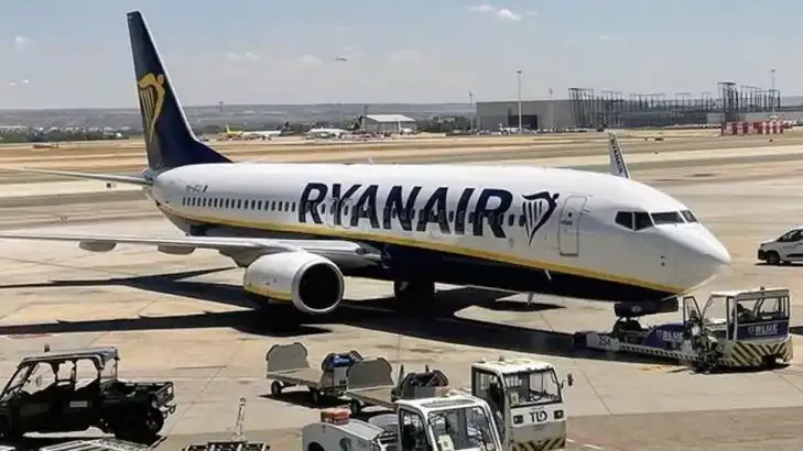 Eine Boeing 737 von Ryanair auf dem Vorfeld von Alghero, mit Terminal und Bodengerät dahinter.