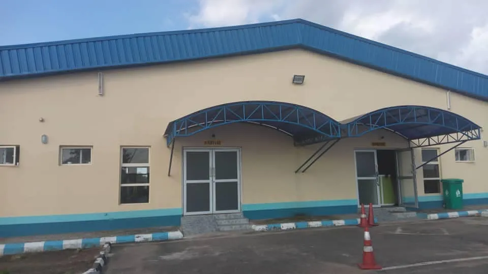 Das Terminal von Akure Airport – cremefarbene Wände unter einem blauen Dach, geschwungene Vordächer über den Eingängen und eine blau-weiße Bordsteinkante.