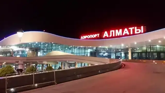 Bei Nacht leuchtet das rote Neon „АЭРОПОРТ АЛМАТЫ“ über der verglasten Front.