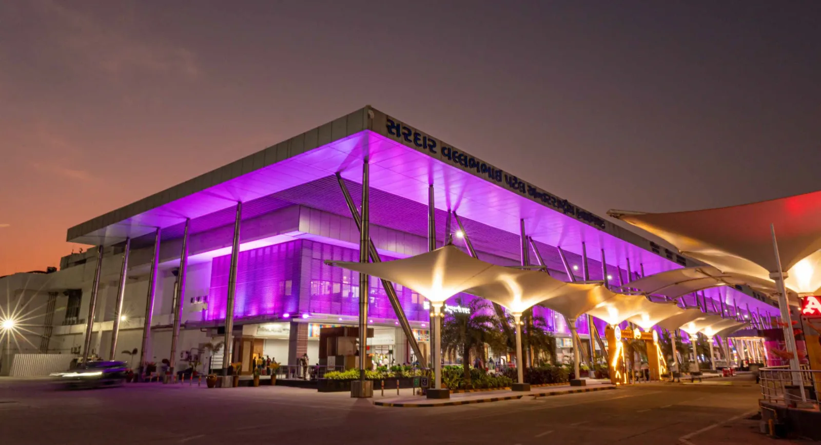 Das Terminal bei Nacht – violettes Licht auf dem auskragenden Dach, Stützen über dem Vorfahrtsbereich und der Flughafenname in Gujarati entlang der Fassade.