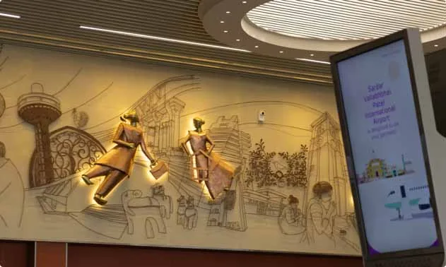 Eine Kunstwand im Terminal – zwei Bronzefiguren vor einer gezeichneten Stadtansicht, mit einem Bildschirm, der den vollständigen Namen des Flughafens zeigt.