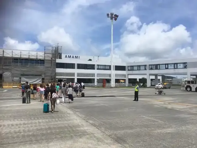 Auf dem Vorfeld vor dem Terminal – Passagiere mit Gepäck auf dem Weg zum Gebäude mit der Aufschrift „AMAMI“, Bodenpersonal weist sie ein.