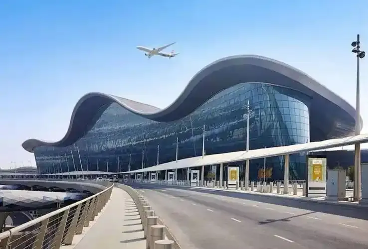 Das Wellendach des Zayed International Airport – ein Verkehrsflugzeug zieht darüber hinweg, im Vordergrund Vorfahrtstraße und Sonnensegel.