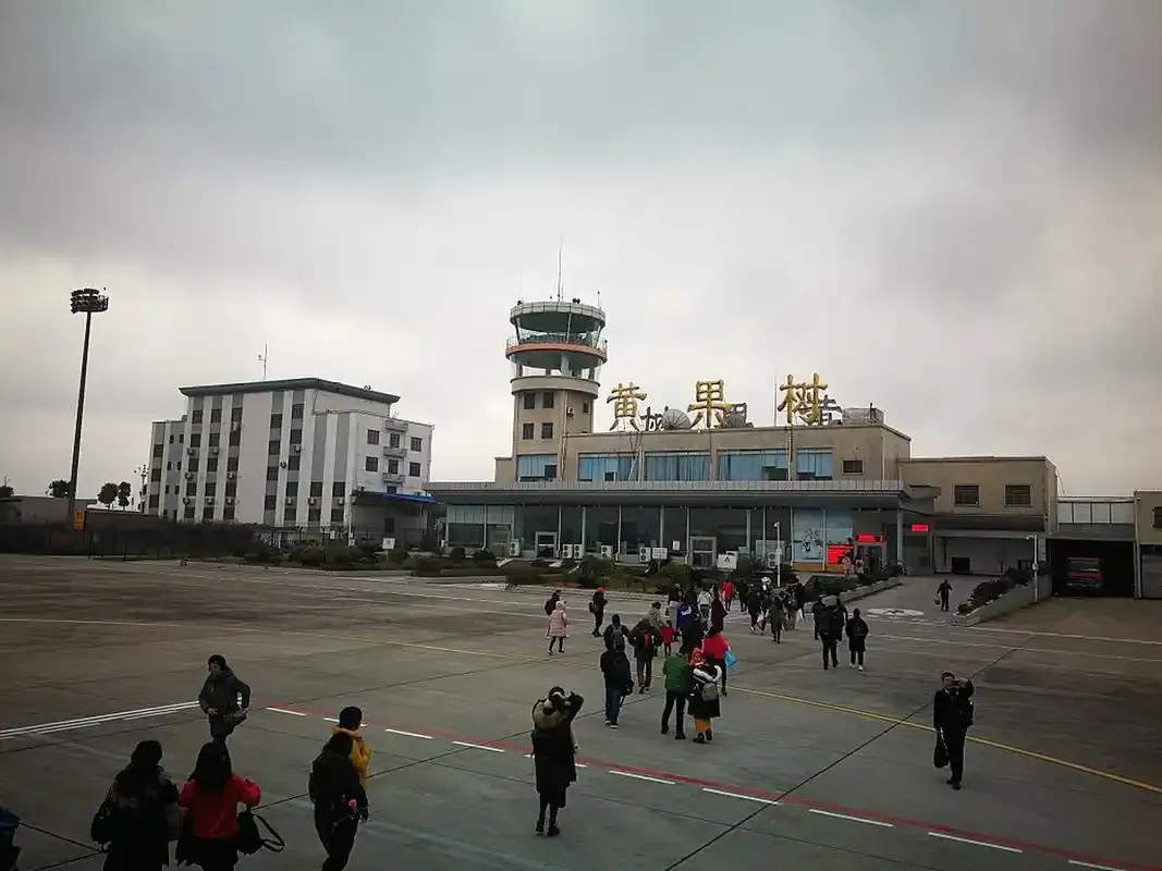 Passagiere gehen zum Einsteigen hinaus – hinter ihnen das vergoldete Schild „黄果树“ und die verglaste Fassade, darüber der Turm.