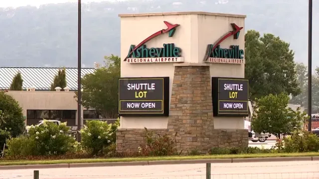 Das Eingangsschild des Asheville Regional Airport – grüne Schrift neben einer Anzeige „SHUTTLE LOT“, dahinter bewaldete Hügel.