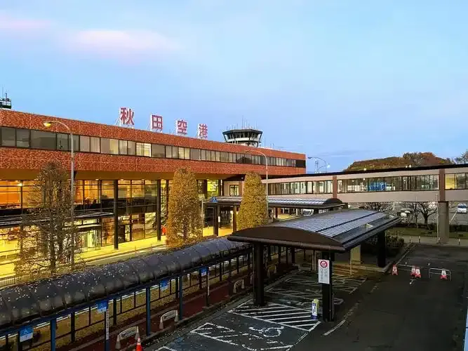 Terminal von Akita in der Dämmerung — „秋田空港“ über der ziegeltonigen Fassade, der Kontrollturm in der Mitte und warmes Licht hinter dem Glas.