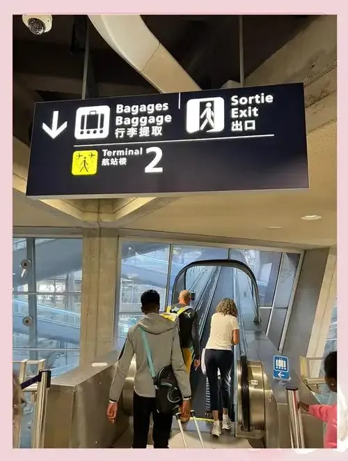 Die Rolltreppe Richtung Terminal 2 – das Schild über dem Kopf zeigt „Bagages / Baggage / Gepäckausgabe“ und „Sortie / Exit“ in drei Sprachen.