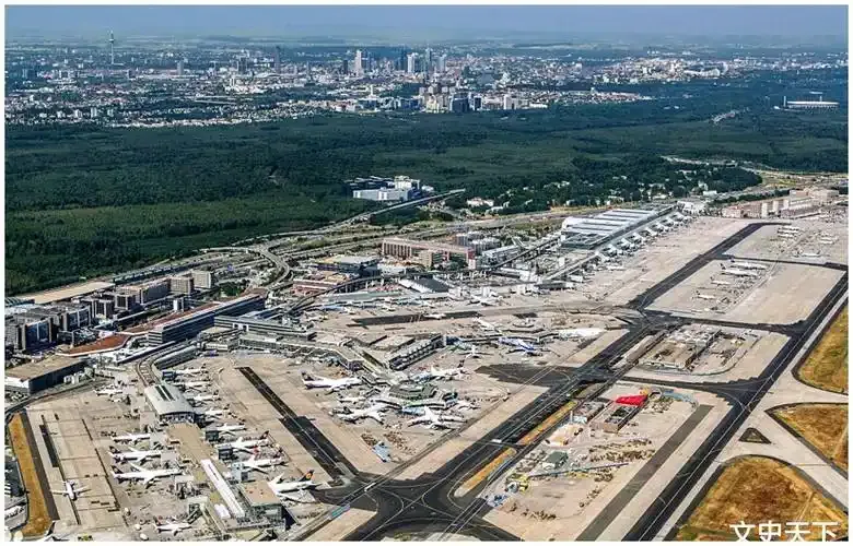 Frankfurt aus der Luft – Flugzeuge und Baufahrzeuge über Vorfeld und Startbahnen verteilt, hinter dem Waldgürtel die Skyline der Stadt.
