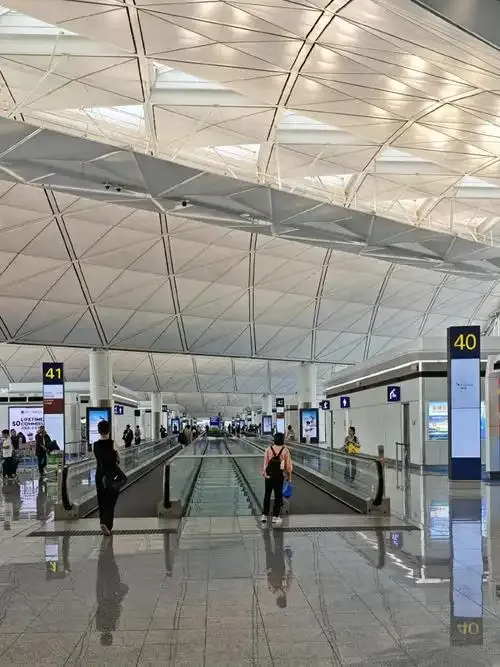 Die Abflugebene von Terminal 1 – Fahrsteige unter der wellenförmigen Decke, darüber die Gate-Schilder 40 und 41, Reisende mit Rollkoffern.