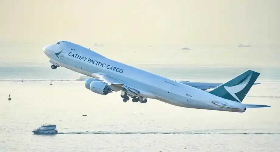 Eine Boeing 747 von Cathay Pacific Cargo hebt von der seeseitigen Startbahn ab, unter dem Flugzeug fährt eine Fähre vorbei.