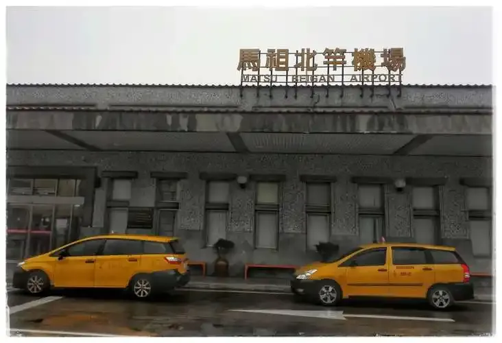 Two yellow taxis wait outside the terminal entrance beneath the 'Matsu Beigan Airport' rooftop sign.
