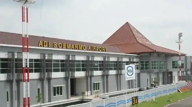 Die Terminalfront – „ADI SOEMARMO AIRPORT“ unter einem Ziegeldach, Fahnenmasten und Zäune in ordentlichen Reihen.